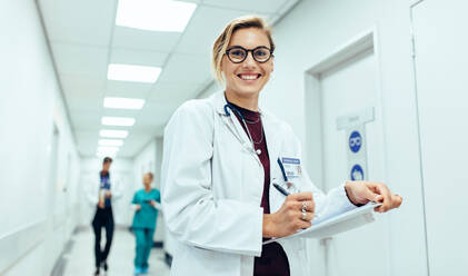Portrait of happy young female physician standing in corridor with clipboard. Caucasian woman working in hospital writing prescription. - JLPSF27568