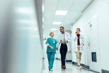 Eine Gruppe von Ärzten mit einem Klemmbrett diskutiert auf dem Krankenhausflur. Ein Arzt und eine Krankenschwester besprechen einen medizinischen Bericht mit einer Kollegin, die mit einem Mobiltelefon spricht. - JLPSF27572