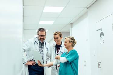 Mature female nurse showing clipboard to doctors in hallway. Team of medics looking at medical report while standing in hospital corridor. - JLPSF27573