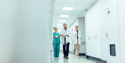 Eine Gruppe von Ärzten mit einem Klemmbrett geht einen Krankenhausflur entlang. Ein Arzt und eine Krankenschwester besprechen einen medizinischen Bericht mit einer Kollegin, die mit einem Mobiltelefon spricht. - JLPSF27594