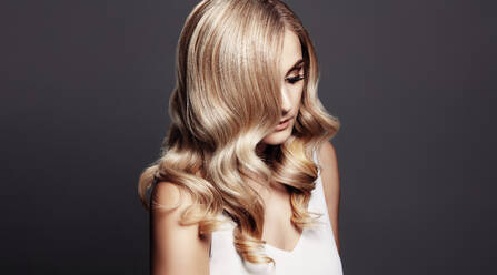 Elegante Frau mit glänzendem, gewelltem blondem Haar. Weibliches Modell mit gewellter Frisur vor grauem Hintergrund. - JLPSF27634