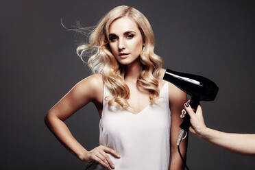 Elegante junge Frau in einem Studio, während die Luft eines Föns ihr Haar bläst. Schöne Blondine mit vom Gebläse geblasenem Haar vor grauem Hintergrund. - JLPSF27636