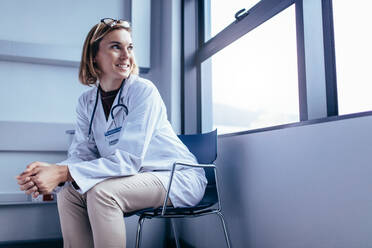 Porträt einer weiblichen medizinischen Fachkraft, die auf einem Stuhl in einem Krankenhauszimmer sitzt und aus dem Fenster schaut. - JLPSF27662