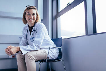 Porträt einer lächelnden Ärztin, die auf einem Stuhl in einem Krankenhauszimmer sitzt und in die Kamera schaut. - JLPSF27663