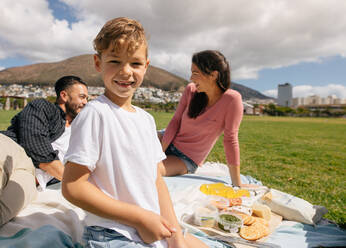 Fröhliches Paar mit ihrem Kind, das einen Ausflug macht. Familie sitzt im Park mit Snacks für ein Picknick. - JLPSF27694