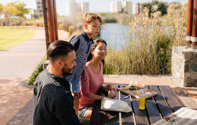 Fröhliches Paar mit ihrem Kind, das einen Ausflug macht. Familie sitzt im Park mit Snacks für ein Picknick. - JLPSF27697