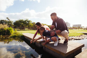 Familie in fröhlicher Stimmung in einem Park. Paar mit Kind sitzt auf der Brücke eines Baches und schaut auf das Wasser. - JLPSF27700