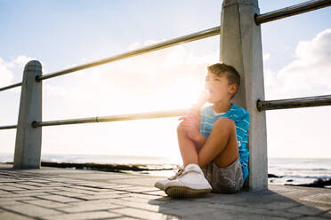 Junge isst ein Eis an einem Geländer am Meer mit der untergehenden Sonne im Hintergrund. Kleiner Junge im Urlaub, der sich ein Eis gönnt. - JLPSF27707
