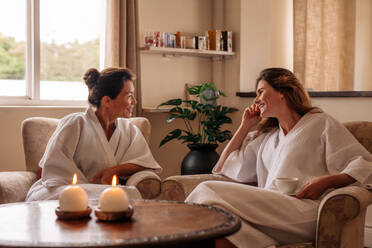 Two female friends chatting while waiting in spa reception area. Women in bathrobe sitting on chairs and talking at health spa. - JLPSF27715