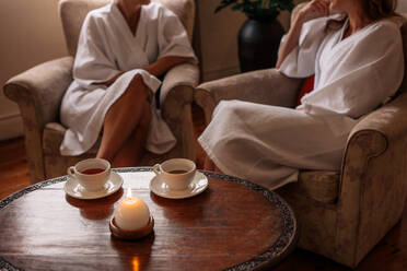 Spa waiting area with women sitting in background. Focus on table with candle and herbal tea cups. - JLPSF27721