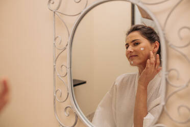 Frau, die im Badezimmer Feuchtigkeitscreme auf ihr Gesicht aufträgt. Reflexion einer Frau, die in den Spiegel schaut und sich mit einer kosmetischen Lotion eincremt. - JLPSF27742