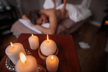 Closeup on candles in front with a relaxed woman getting back massage in background at health spa. Woman receiving massage in spa salon with focus on candlelight on table. - JLPSF27748