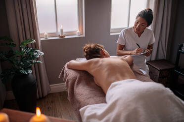Female massage therapist talking to woman at wellness center and making notes. Beautician examining female client before spa treatment. - JLPSF27752