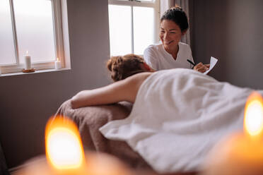 Beautician examining female client before spa treatment and making notes for the therapy. Female massage therapist talking to woman at wellness center and making notes. - JLPSF27755