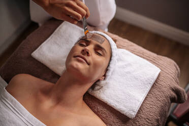 Frau auf Massagetisch liegend mit Kosmetikerin, die eine Gesichtsmaske aufträgt. Frau erhält eine Gesichtsmaskenbehandlung im Schönheitssalon. - JLPSF27762