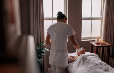 Beautician applying herbal face mask to female lying on massage table in beauty spa. Female performing a beauty treatment on woman client at health spa. - JLPSF27770