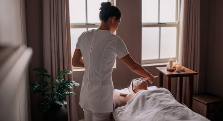 Female applying facial mask to woman face in health spa. Beautician herbal face mask to female lying on massage table in beauty spa. - JLPSF27771