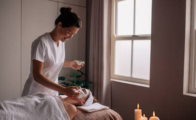 Young woman having spa procedure on her face. Woman lying on spa table and cosmetologist applying cosmetic mask on the her face in sap salon. - JLPSF27772