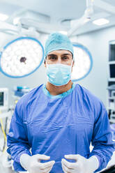 Porträt eines Chirurgen mit OP-Handschuhen und Kittel im Operationssaal. Arzt in Kittel und medizinischer Maske im Operationssaal eines Krankenhauses. - JLPSF27793