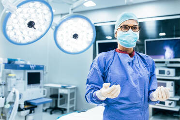 Portrait of female surgeon in surgical uniform in operation theater looking away. Doctor in scrubs and medical mask in modern hospital operating room. - JLPSF27797