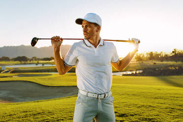 Aufnahme eines männlichen Golfspielers mit Golfschläger auf einem Golfplatz. Ein junger Mann hält einen Golfschläger und schaut an einem sonnigen Tag weg. - JLPSF27818