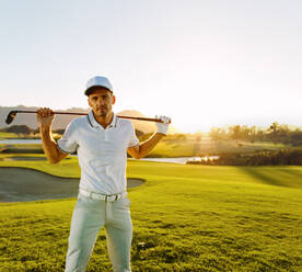 Porträt eines jungen Mannes, der an einem sonnigen Tag einen Golfschläger hält und in die Kamera schaut. Professioneller männlicher Golfer mit Golfschläger auf dem Platz. - JLPSF27819