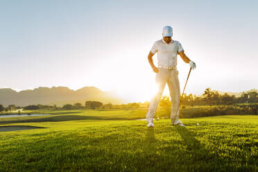 Junger Mann in voller Länge auf einem Golfplatz bei Sonnenuntergang, männlicher Profigolfer mit Golfschläger in der Hand. - JLPSF27820