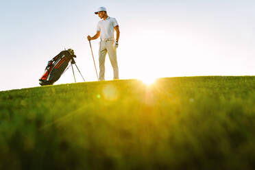 Golfspieler in voller Länge, der seine Tasche betrachtet, während er an einem sonnigen Tag auf dem Feld steht. Niedrigwinkelaufnahme eines männlichen Golfspielers auf dem Golfplatz. - JLPSF27822