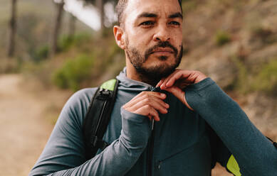 Porträt eines fitten jungen Mannes im Freien auf einem Bergpfad. Gesunder junger Läufer, der auf einer Landstraße den Reißverschluss seiner Jacke zurechtrückt. - JLPSF27830