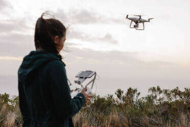 Rear view shot of young woman watching and navigating a flying drone in sky over countryside. - JLPSF27842
