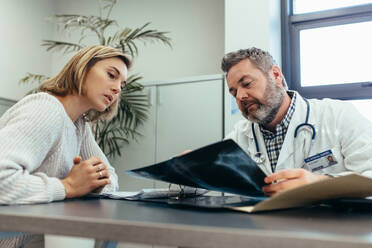 Ein Arzt zeigt einer Frau, die am Schreibtisch sitzt, die Diagnose eines Röntgenbildes. Ein Arzt hält die Ergebnisse eines medizinischen Scans und diskutiert mit einer Patientin. - JLPSF27846