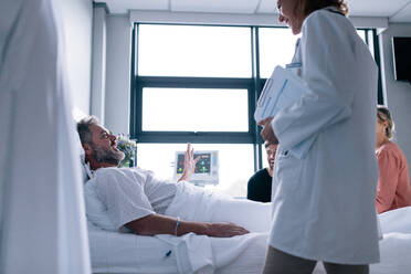 Eine Ärztin besucht einen Patienten in einem Krankenhauszimmer, ein kranker Mann liegt im Bett, seine Familie sitzt neben ihm und der Arzt. - JLPSF27852