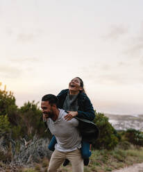 Smiling young man carrying his girlfriend on back. Couple enjoying piggyback ride in countryside. - JLPSF27859