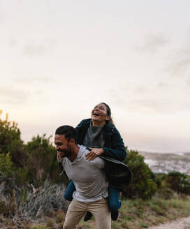 Lächelnder junger Mann, der seine Freundin auf dem Rücken trägt. Paar genießt Huckepackfahrt auf dem Lande. - JLPSF27859
