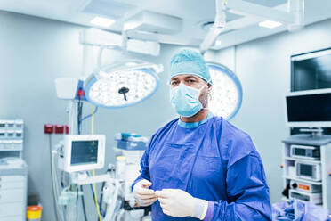 Porträt eines männlichen Chirurgen im Operationssaal, der in die Kamera schaut, Arzt in Kittel und medizinischer Maske in einem modernen Krankenhaus. - JLPSF27860