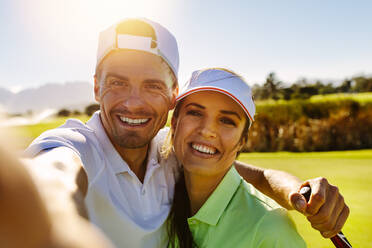 Lächelnde männliche und weibliche Golfer, die ein Selfie auf einem Feld an einem sonnigen Tag machen. Glückliches junges Paar, das ein Selbstporträt auf einem Golfplatz macht. - JLPSF27861