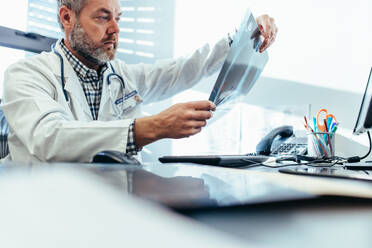 Älterer Arzt, der an seinem Schreibtisch Röntgenbilder betrachtet. Männlicher Arzt, der in seinem Büro sitzt und einen medizinischen Scan untersucht. - JLPSF27878