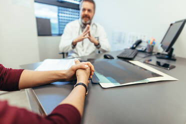 Frau sitzt an einem Tisch in einer Klinik mit einem Arzt im Hintergrund. Fokus auf die Hände der Patientin, die den Arzt aufsucht. Frau trifft sich mit einem Arzt, um sich beraten zu lassen. - JLPSF27882