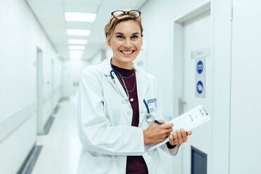 Porträt einer glücklichen jungen Ärztin, die in einem Krankenhauskorridor steht und einen medizinischen Bericht auf einem Klemmbrett schreibt. - JLPSF27914