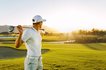 Aufnahme eines männlichen Golfspielers mit Golfschläger auf einem Golfplatz. Ein junger Mann hält einen Golfschläger und schaut an einem sonnigen Tag weg. - JLPSF27922