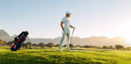 Ein junger Mann, der bei Sonnenuntergang auf einem Golfplatz steht und wegschaut, hält einen Golfschläger auf einem Feld. - JLPSF27930