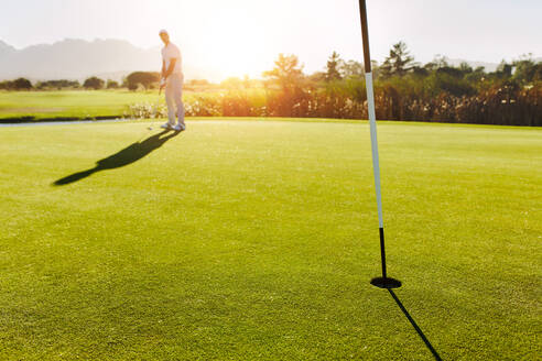 Golfloch und Flagge im grünen Feld mit Spieler im Hintergrund. Professioneller männlicher Golfer, der den Ball schlägt. - JLPSF27944