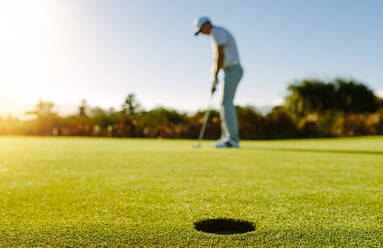 Golfspieler beim Einlochen des Balls. Horizontale Aufnahme eines Golflochs im grünen Feld mit Golfer im Hintergrund. - JLPSF27947