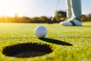Professioneller Golfer beim Einlochen des Balls. Golfball am Rand des Lochs mit Spieler im Hintergrund an einem sonnigen Tag. - JLPSF27949