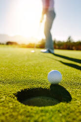 Golfball am Rande des Lochs mit Spieler im Hintergrund. Professioneller Golfer, der den Ball an einem sonnigen Tag ins Loch schlägt. - JLPSF27950