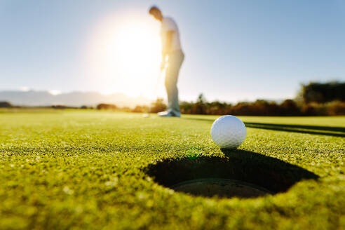 Profigolfer beim Einlochen des Golfballs. Golfball am Loch mit Spieler im Hintergrund an einem sonnigen Tag. - JLPSF27951