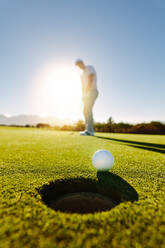 Vertical shot of professional golfer putting golf ball in to the hole. Golf ball by the hole with player in background on a sunny day. - JLPSF27952