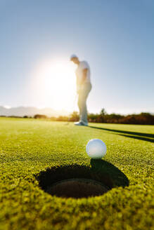 Vertikale Aufnahme eines professionellen Golfspielers, der einen Golfball in das Loch schlägt, mit einem Spieler im Hintergrund an einem sonnigen Tag. - JLPSF27952