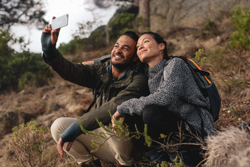 Verliebtes junges Paar, das auf einem Bergpfad sitzt und ein Selfie macht. Glücklicher junger Mann und Frau, die auf dem Lande wandern und sich mit einem Mobiltelefon unterhalten. - JLPSF28004