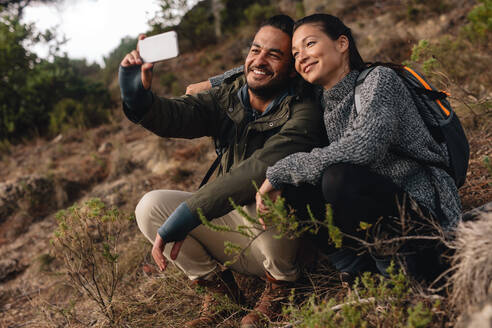 Verliebtes Paar, das auf einem Bergpfad sitzt und ein Selfie macht. Junger Mann und Frau wandern auf dem Land und sprechen Selbstporträt mit Handy. - JLPSF28005